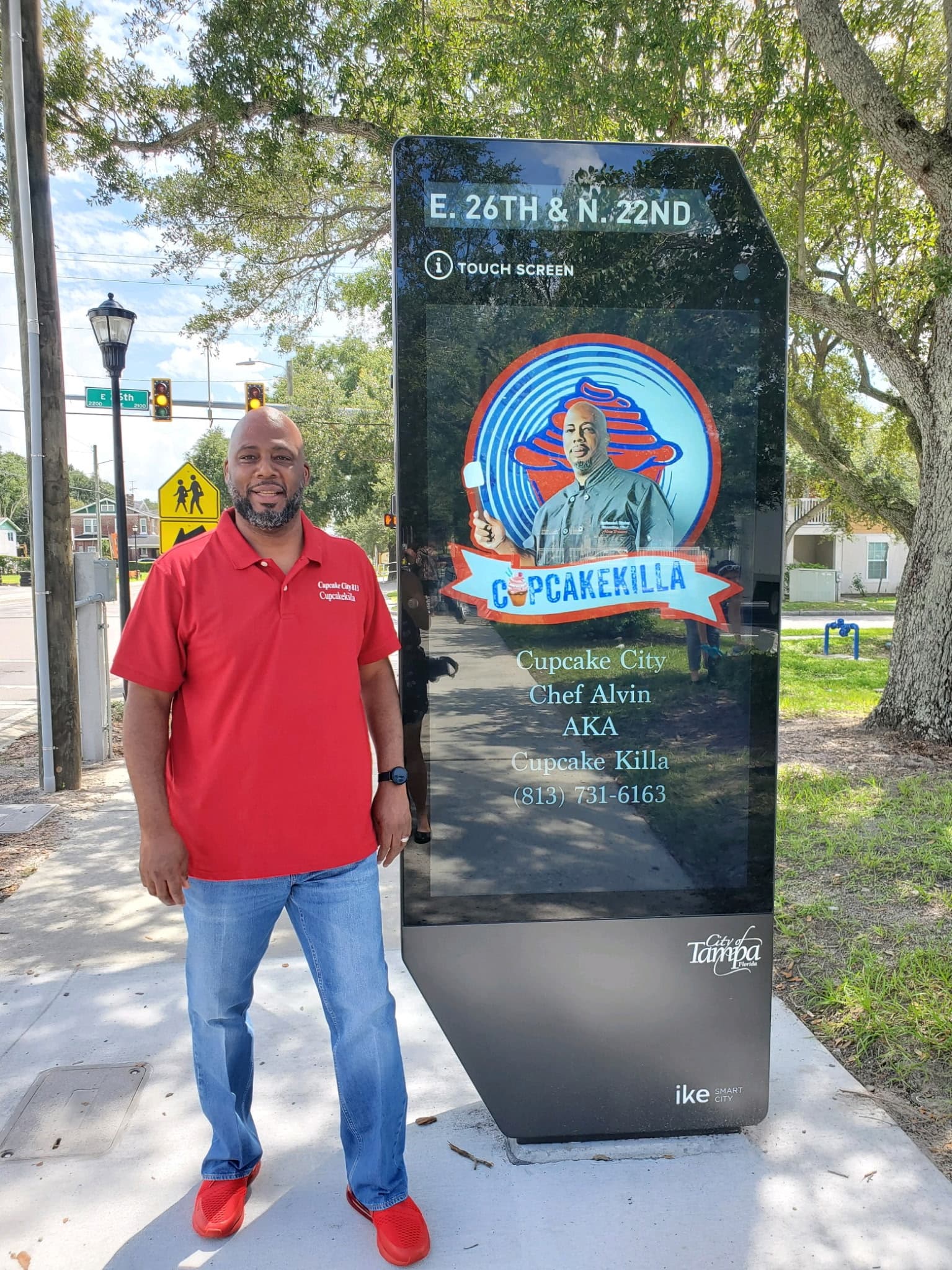 Chef Alvin standing beside a City of Tampa smart kiosk featuring his Cupcakekilla logo and Cupcake City phone number
