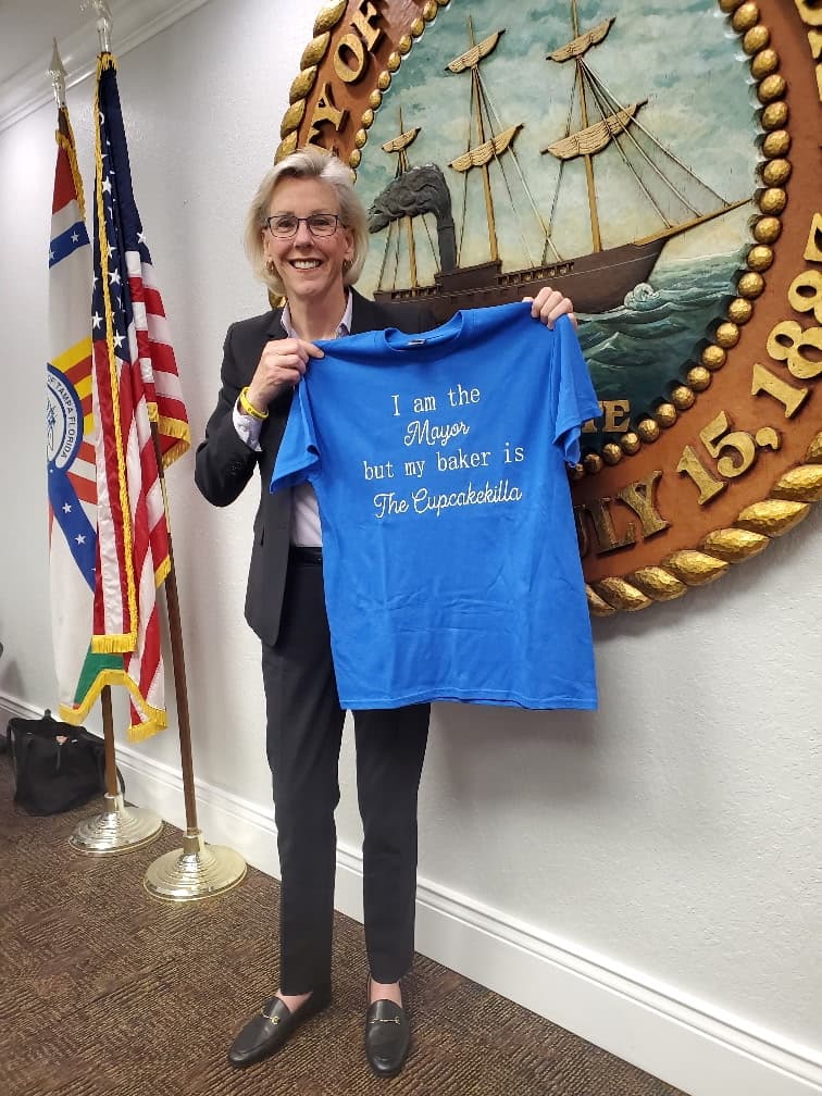 Tampa Mayor Jane Castor holding up a blue shirt that reads: I am the Mayor but my baker is The Cupcakekilla